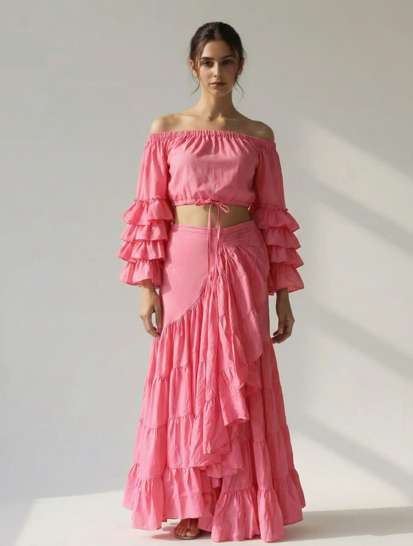 Model standing in a front pose wearing a pink cotton co-ord set, featuring an off-shoulder crop top with tiered ruffle sleeves and a matching wrap-around maxi skirt with a waist tie.