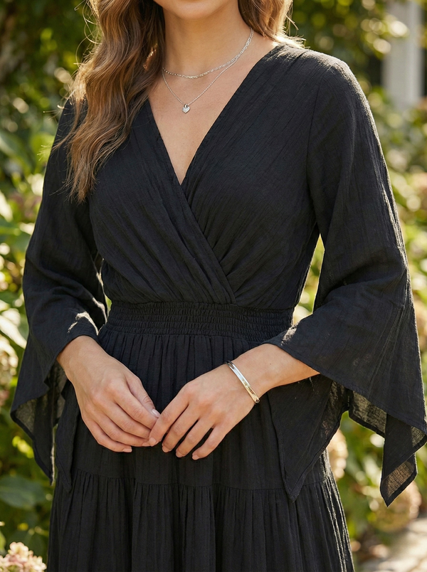 Close up view of Model standing on a stone path wearing a black, floor-length pure cotton maxi dress with a V-neckline and gathered tiered skirt.