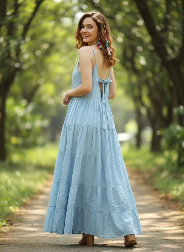 Woman in sky serenity light blue tiered maxi dress, looking back and smiling, showing back tie detail, on pathway