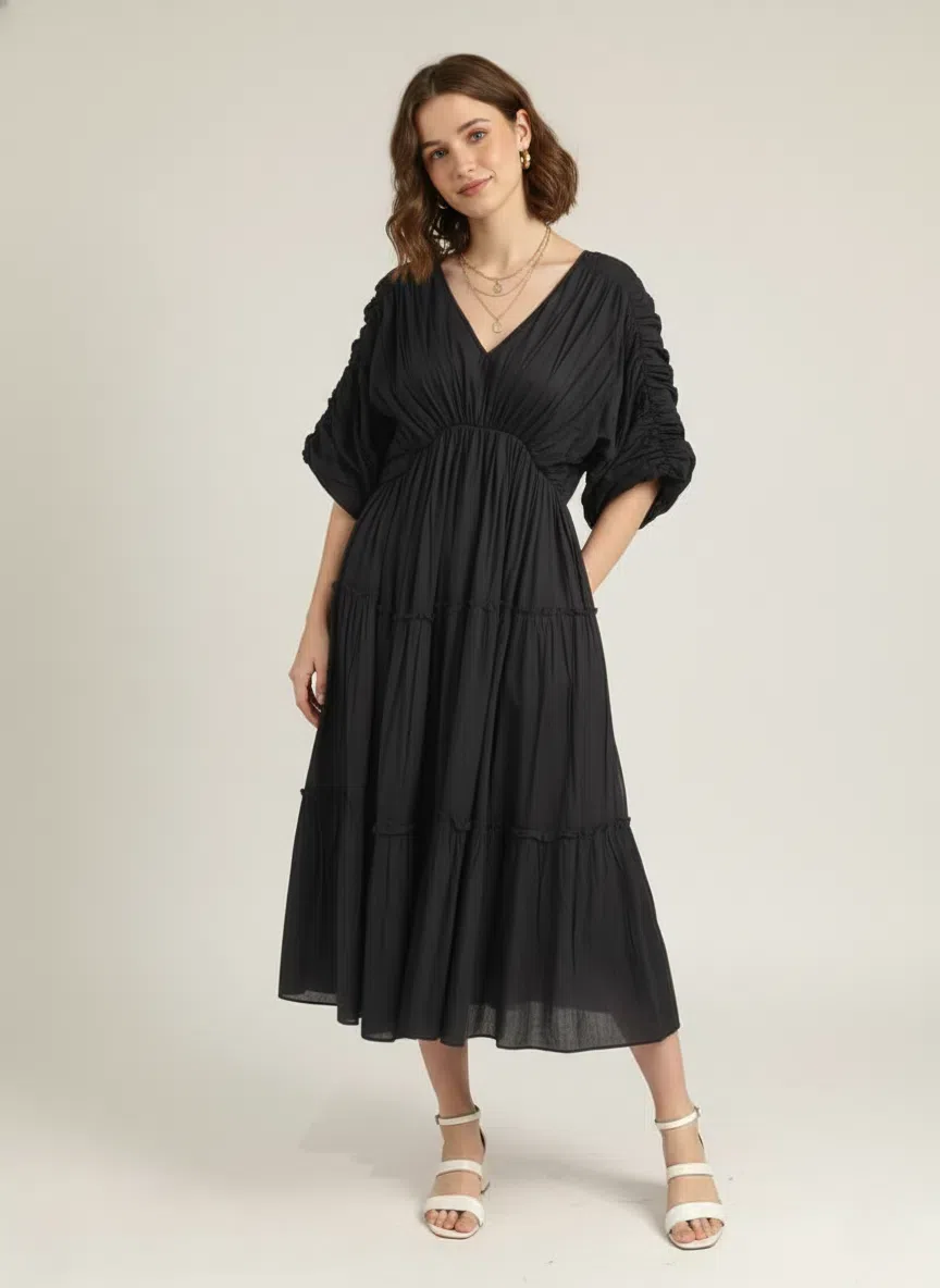 Front view of a woman standing, looking straight at the camera, wearing a classic black pure cotton tiered midi dress with a V-neck and ruched short sleeves, against a plain white background.