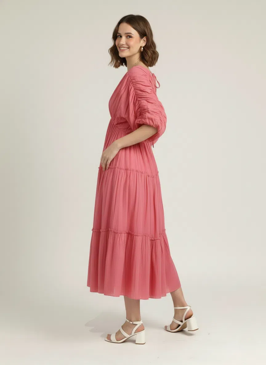 Side profile of a woman smiling over her shoulder, wearing a dusty rose pink pure cotton tiered midi dress with ruched sleeves, against a plain white background.