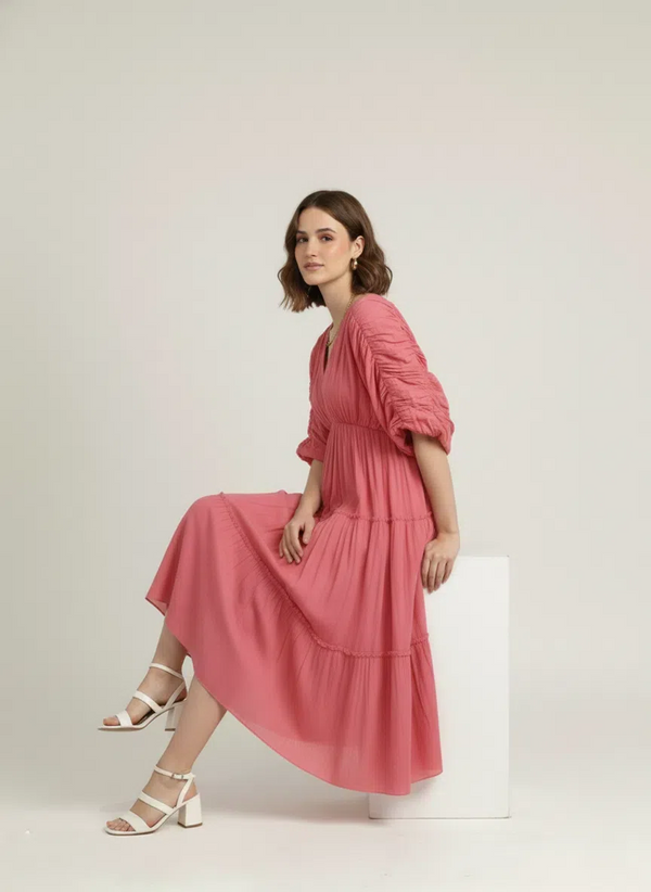 A woman sits elegantly on a white block, wearing a dusty rose pink pure cotton tiered midi dress, with the skirt draped gracefully, against a plain white background.