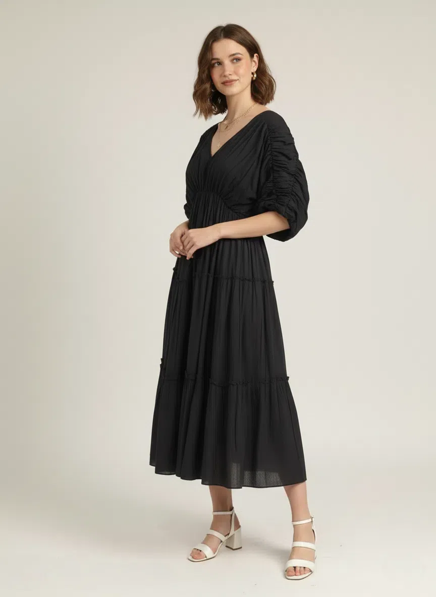 Side profile of a woman wearing a classic black pure cotton tiered midi dress with ruched sleeves and a V-neck, looking over her shoulder against a plain white background.