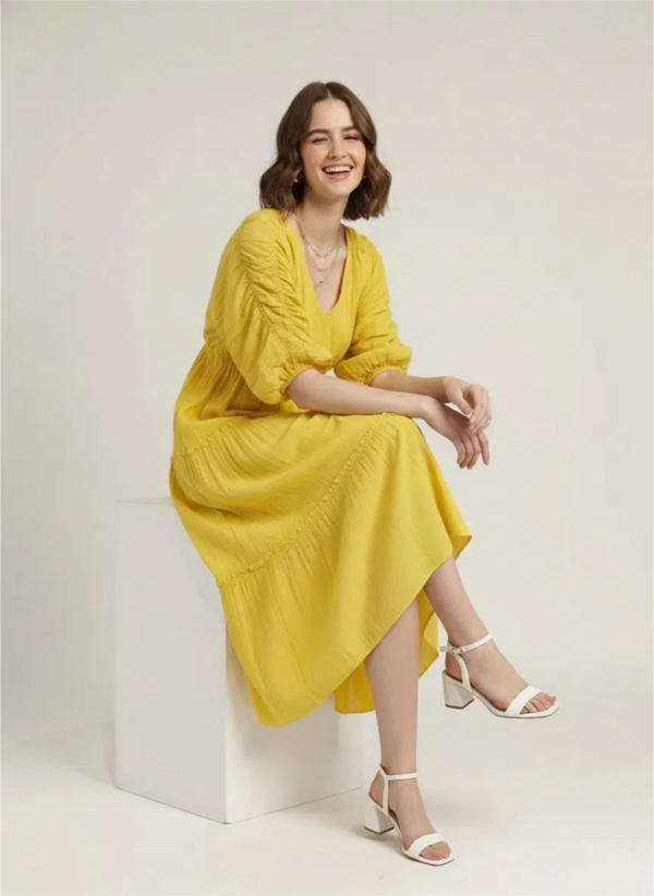 A woman sits casually on a white block, winking and smiling, wearing a luminous lemon yellow pure cotton tiered midi dress with ruched sleeves, against a plain white background.