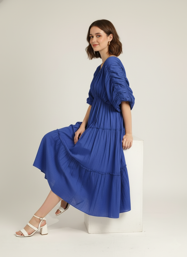 A woman sits gracefully on a white block, smiling, wearing a vivid cerulean blue pure cotton tiered midi dress with ruched sleeves, against a plain white background.