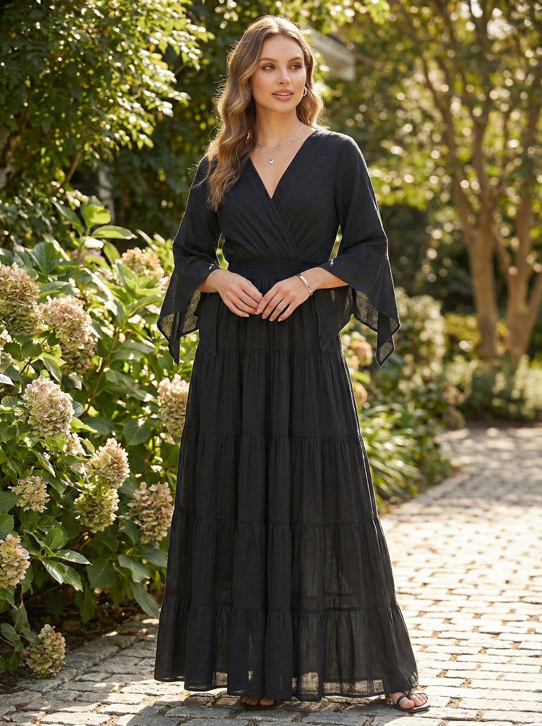 Model standing on a stone path wearing a black, floor-length pure cotton maxi dress with a V-neckline and gathered tiered skirt.