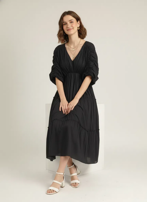 A woman sits gracefully on a white block, smiling, wearing a classic black pure cotton tiered midi dress with a V-neck and ruched sleeves, against a plain white background.