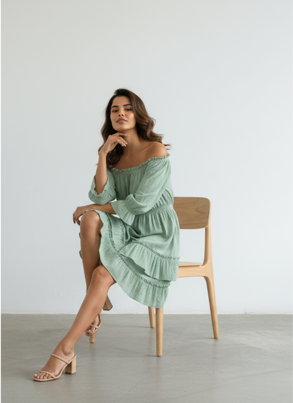 A woman sits gracefully on a wooden chair, wearing a serene sage green pure cotton off-shoulder tiered mini dress, with one hand on her chin, against a plain white background