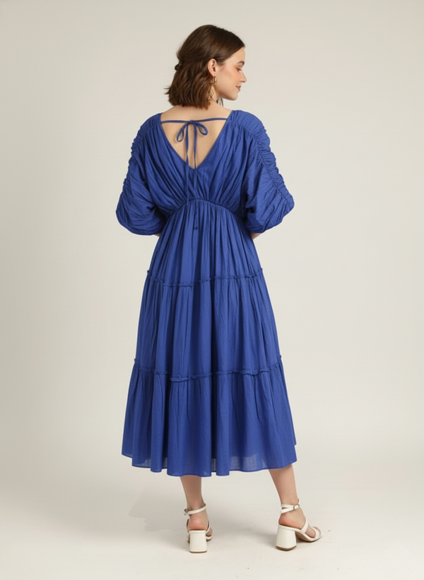 Back view of a woman wearing a vivid cerulean blue pure cotton tiered midi dress with ruched sleeves and a V-back tie detail, against a plain white background.