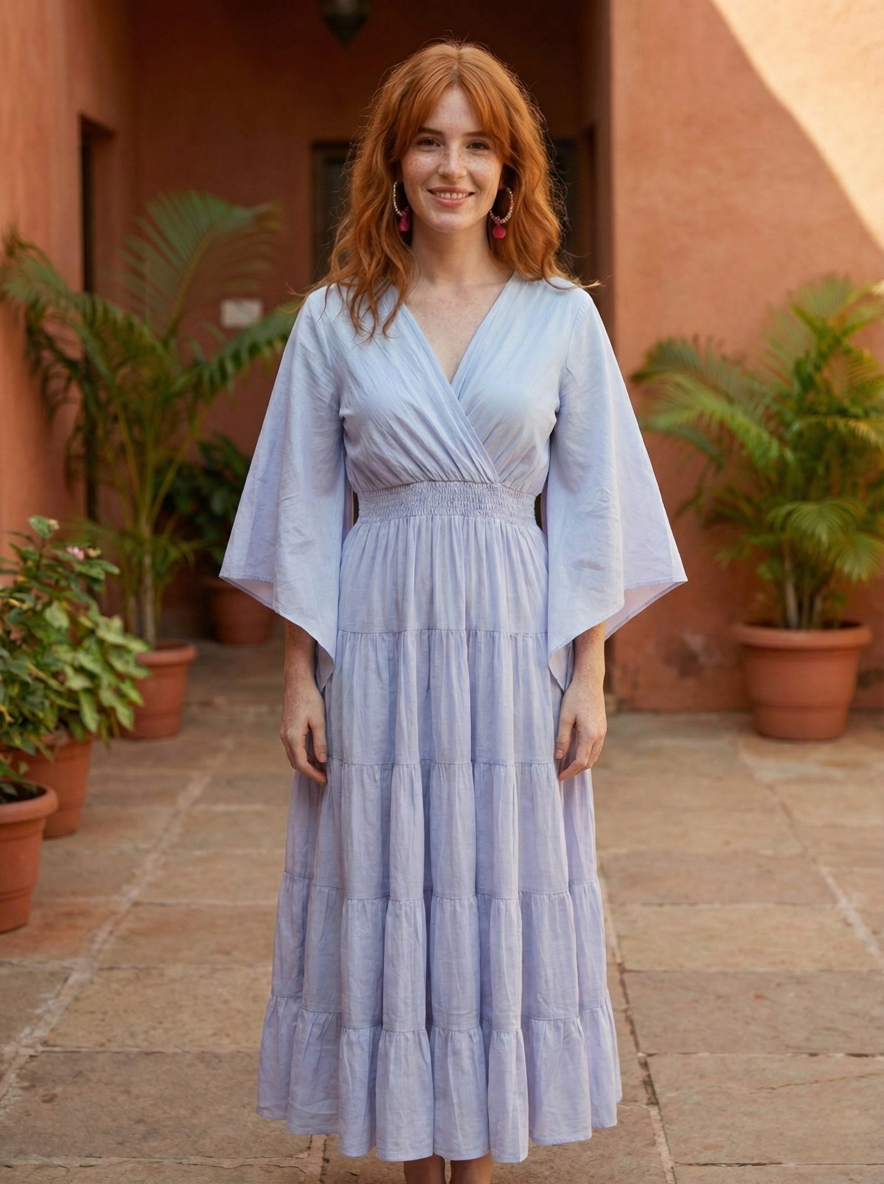 Woman in a light lavender kimono style maxi dress standing in front of a building with plants.