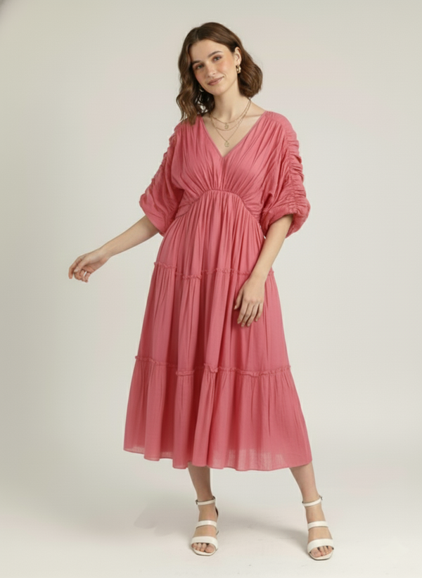 A woman stands, wearing a dusty rose pink pure cotton tiered midi dress with a V-neck and ruched short sleeves, against a plain white background.
